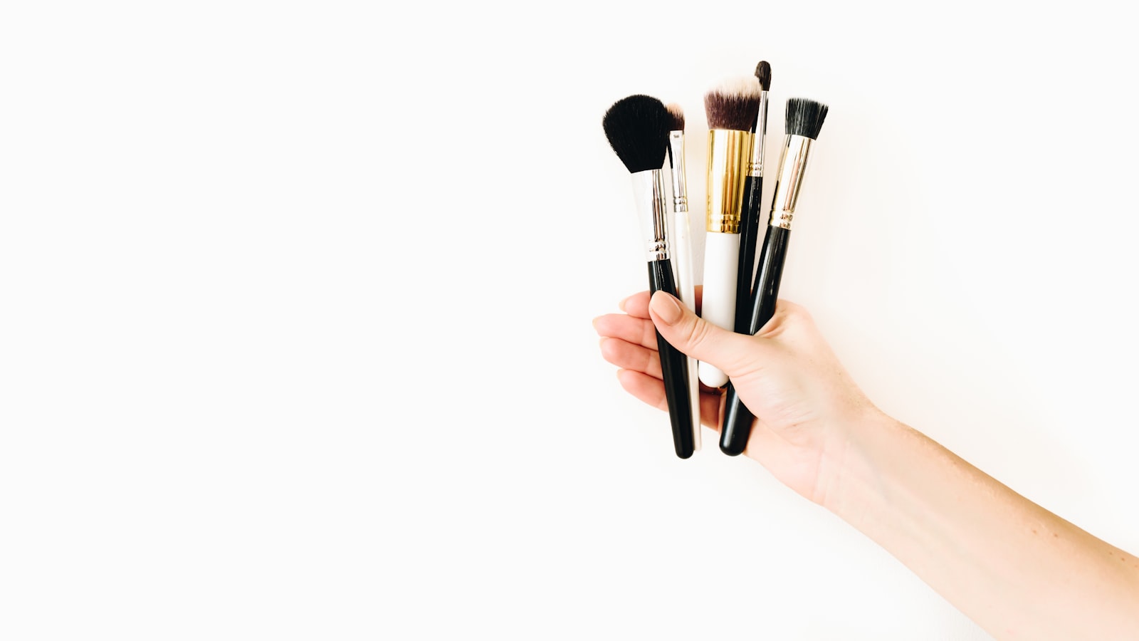 A hand holds five cosmetic brushes against a white background