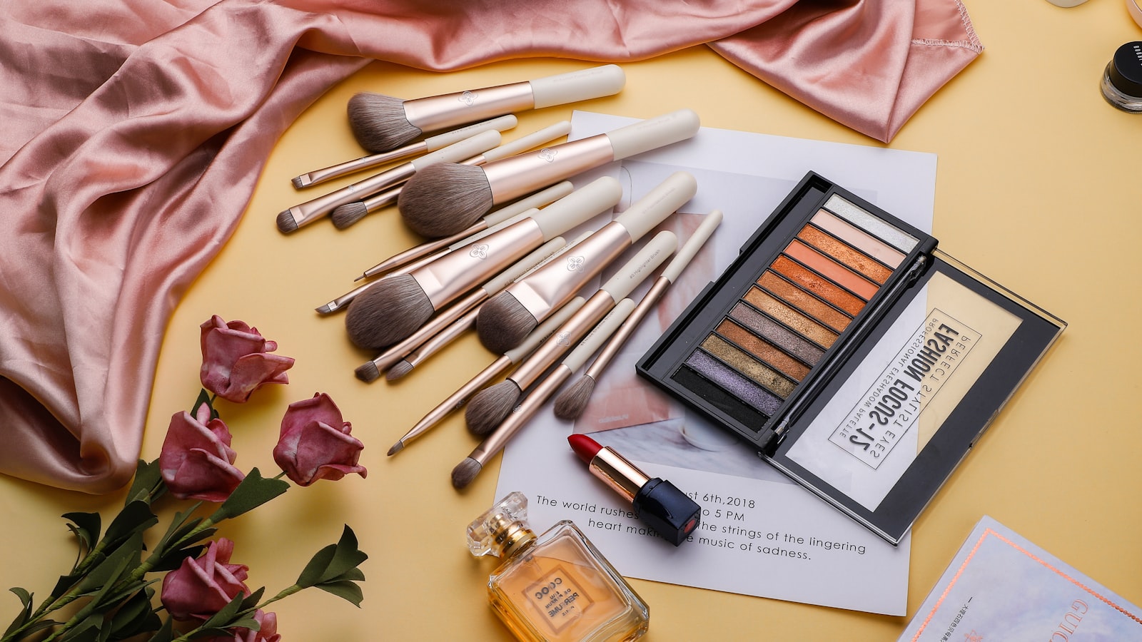 Makeup brushes, an eyeshadow palette, lipstick, and perfume are laid on a yellow surface with pink fabric and paper