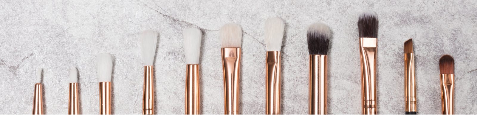 A row of ten makeup brushes with rose gold ferrules on a gray marble surface