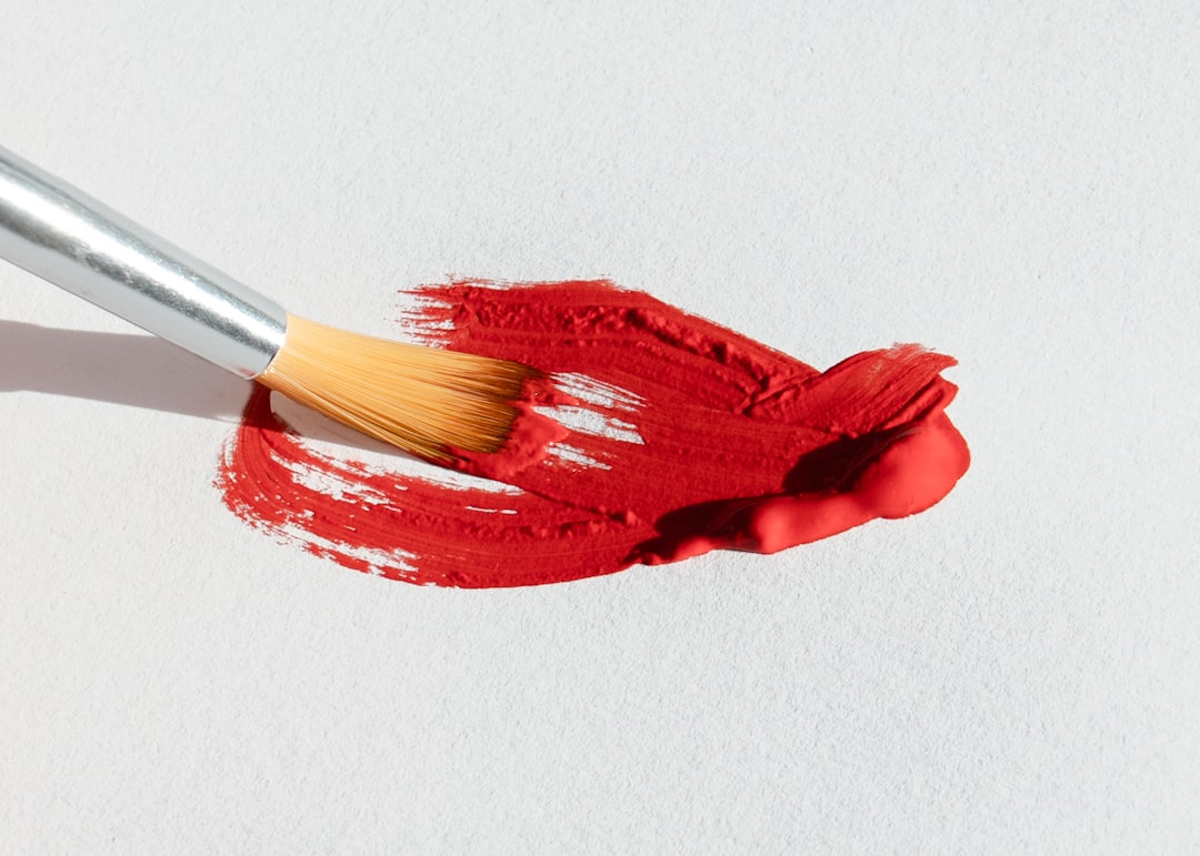 A paintbrush pulls a thick stroke of red paint across a textured white surface