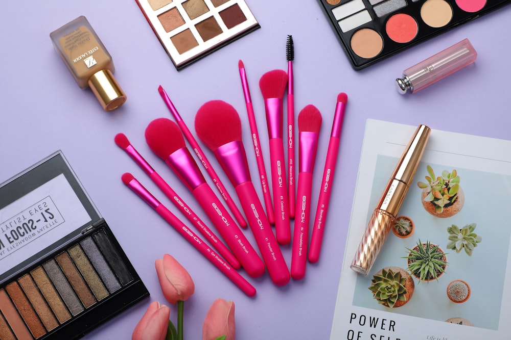 A collection of makeup items and pink-handled brushes is arranged on a purple surface
