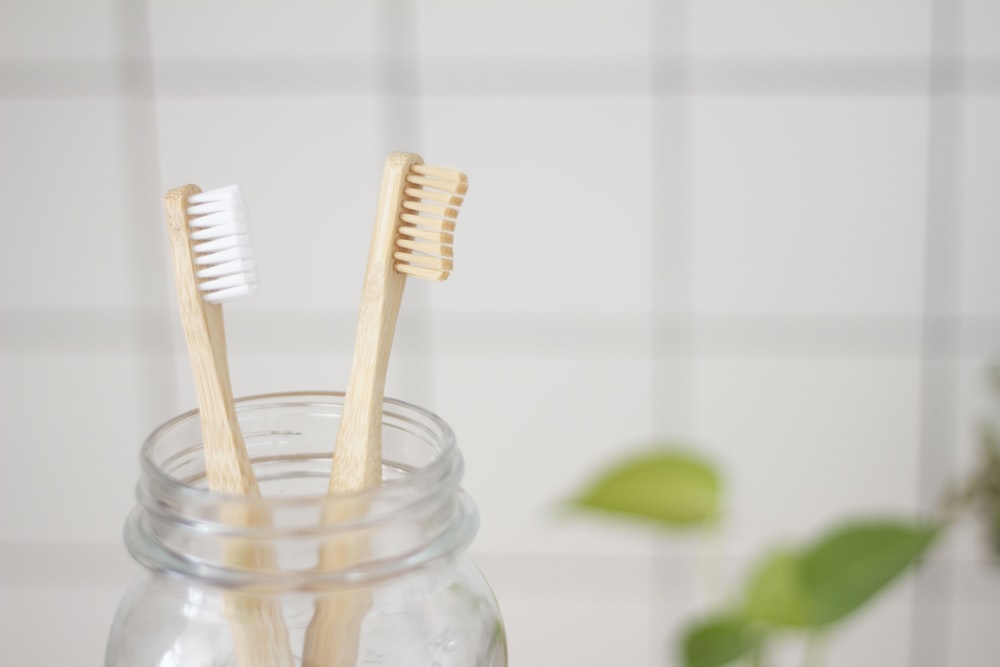 Two wooden toothbrushes in a glass jar against a white tile wall