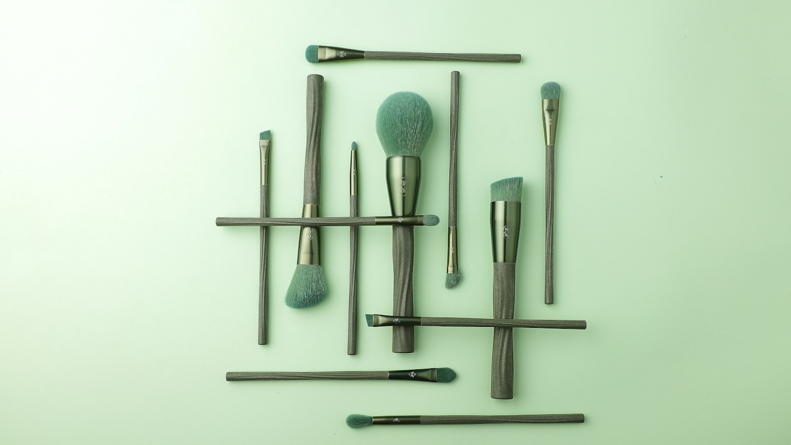 Overhead shot of ten sage green cosmetic brushes arranged on a pale green surface