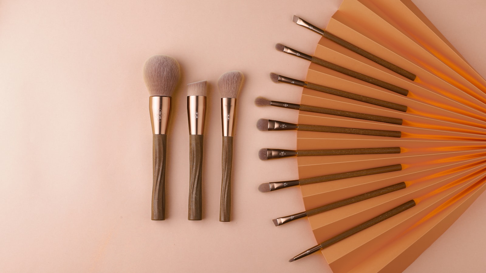 Twelve makeup brushes are arranged on a pink surface next to an orange paper fan