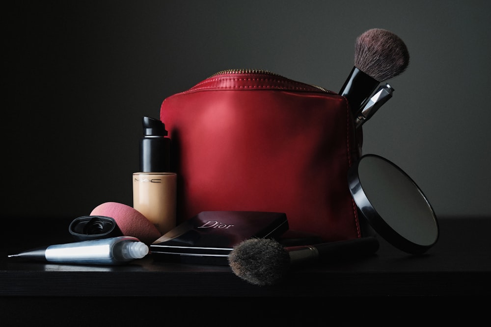 A red makeup bag with brushes, foundation, and a compact mirror on a dark surface