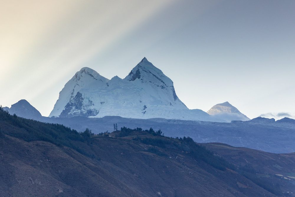Sunlight slants across a snow-capped mountain range rising above dark forested hills