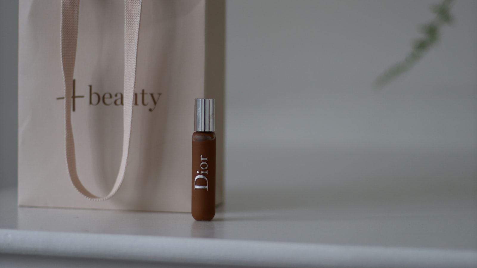 A small Dior vial with a silver cap sits in front of a light pink bag