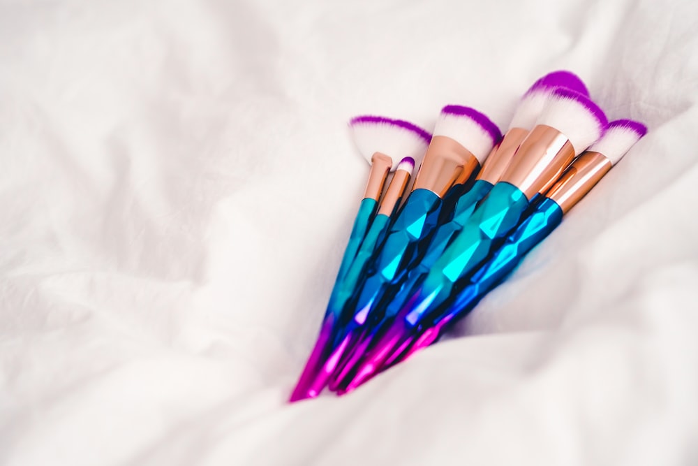 A cluster of cosmetic brushes with turquoise and purple gradient handles rests on a white sheet