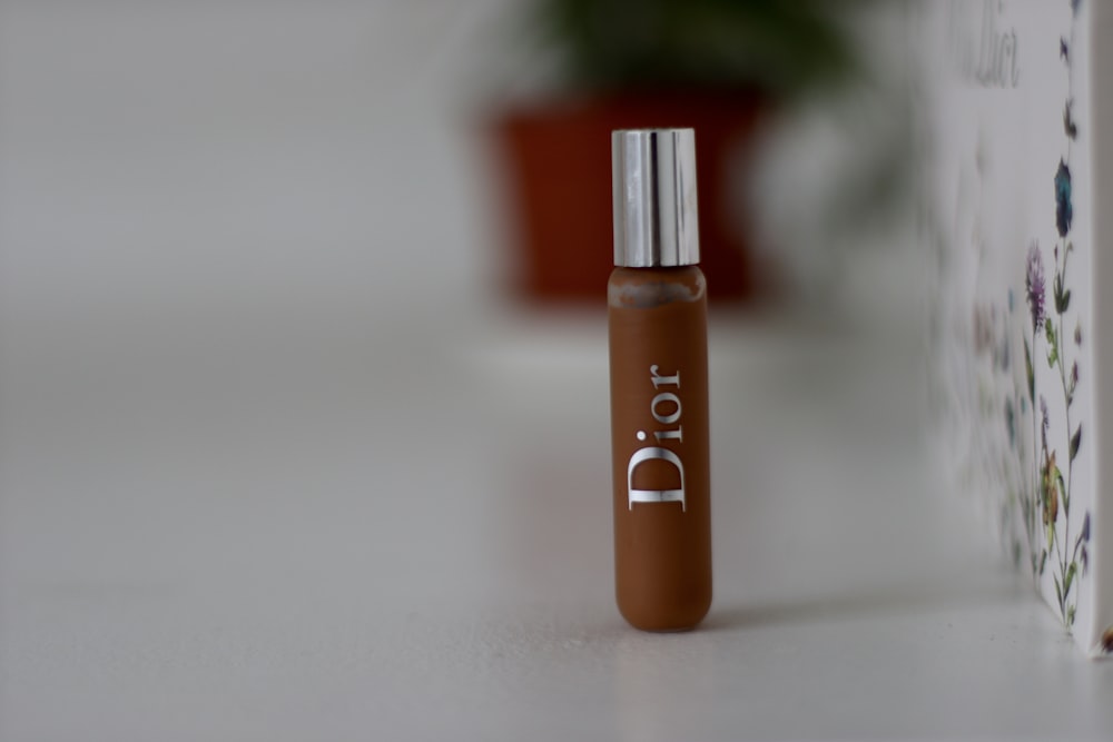 A small brown Dior vial stands upright on a white surface with a blurred background