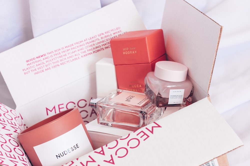 An open cardboard box contains cosmetics, a candle, and a perfume bottle