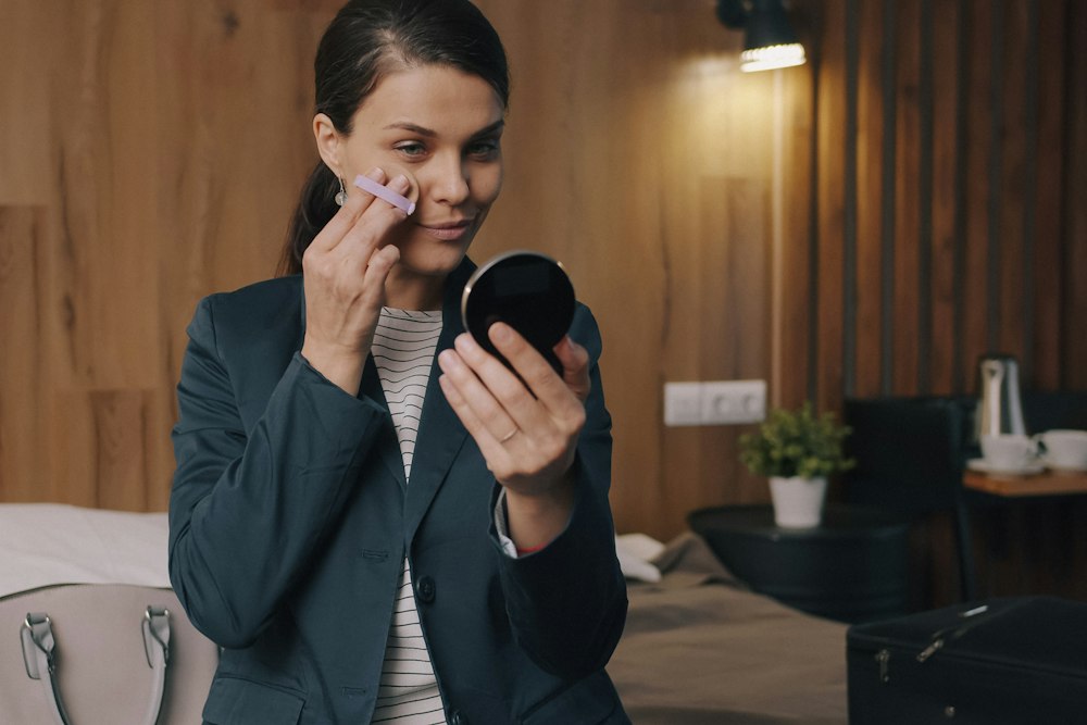 A woman in a blazer holds a small mirror and applies powder to her cheek with a sponge
