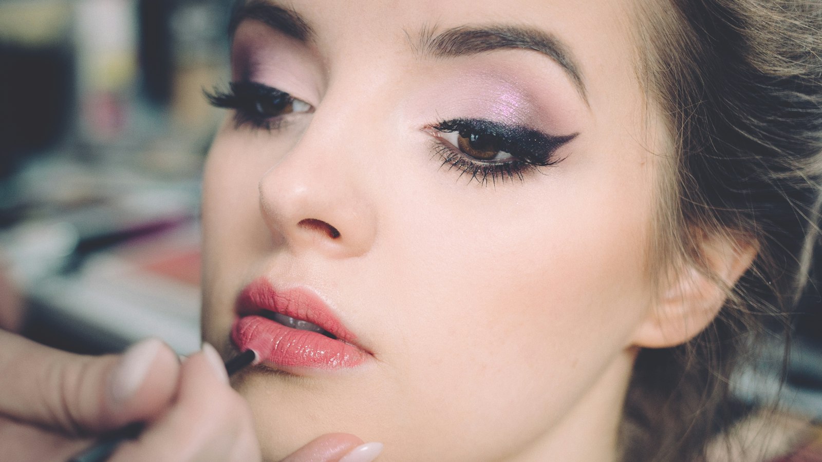 Close-up of a hand applying lipstick to a woman's lips, with eye makeup visible