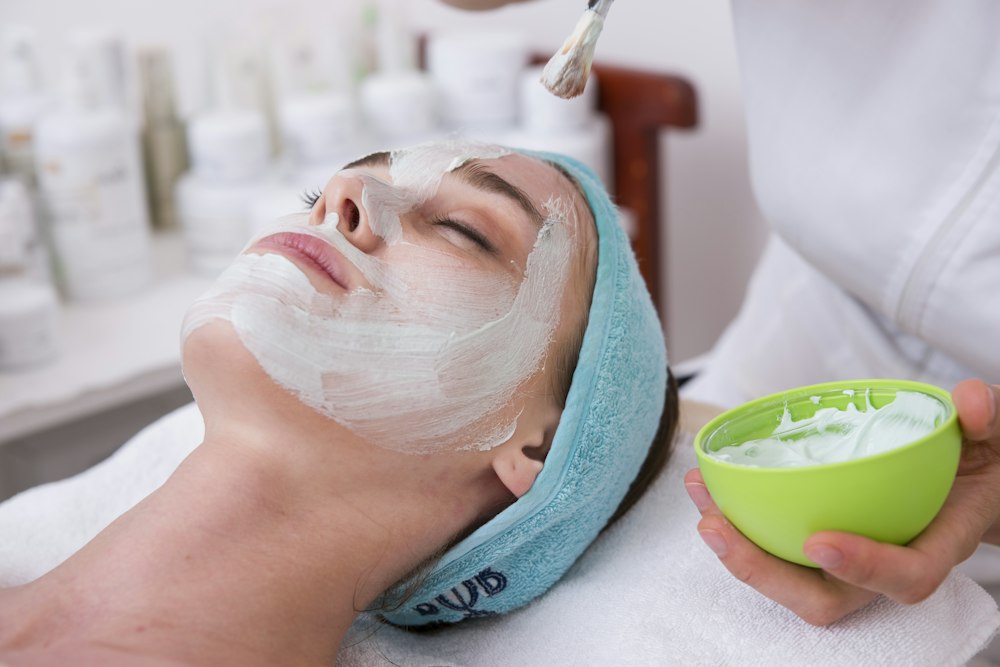 A person applies cream to a reclining woman's face using a brush and green bowl