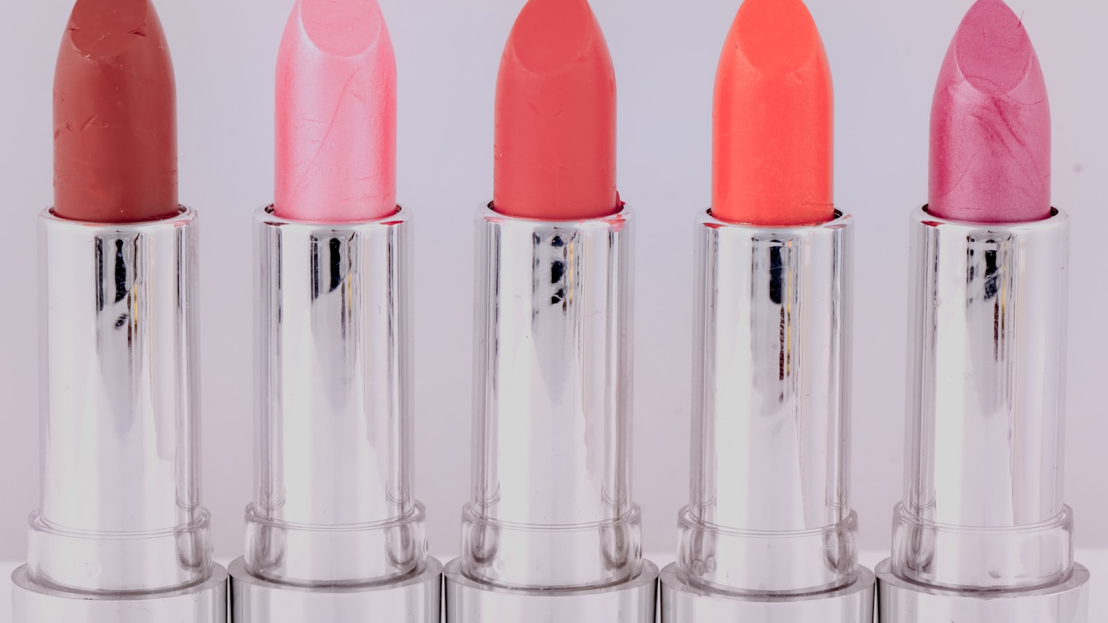 Five open lipsticks in silver tubes stand in a row against a white background
