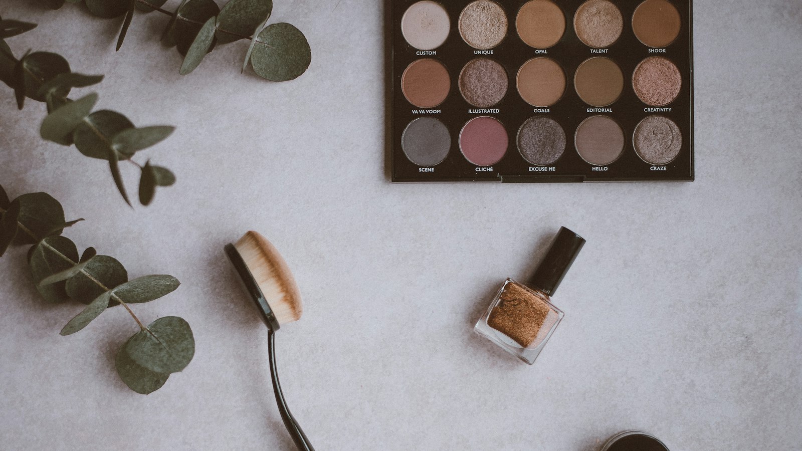 An eyeshadow palette, brush, and bottle of polish lie beside eucalyptus sprigs