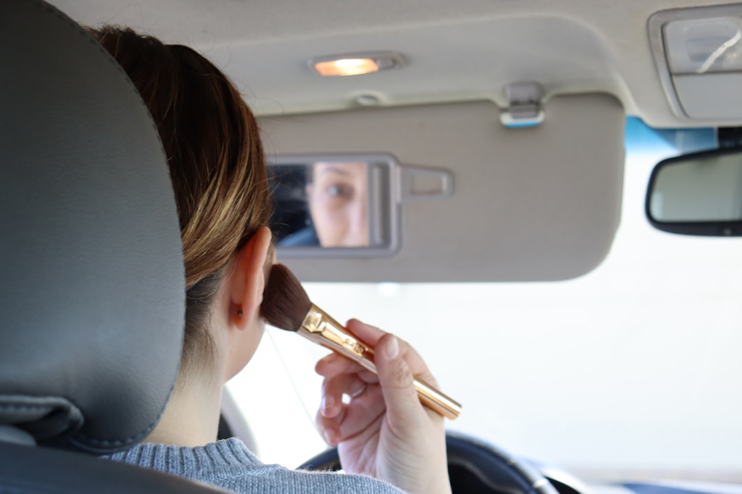 A person in a car applies makeup using a brush, reflected in the visor mirror