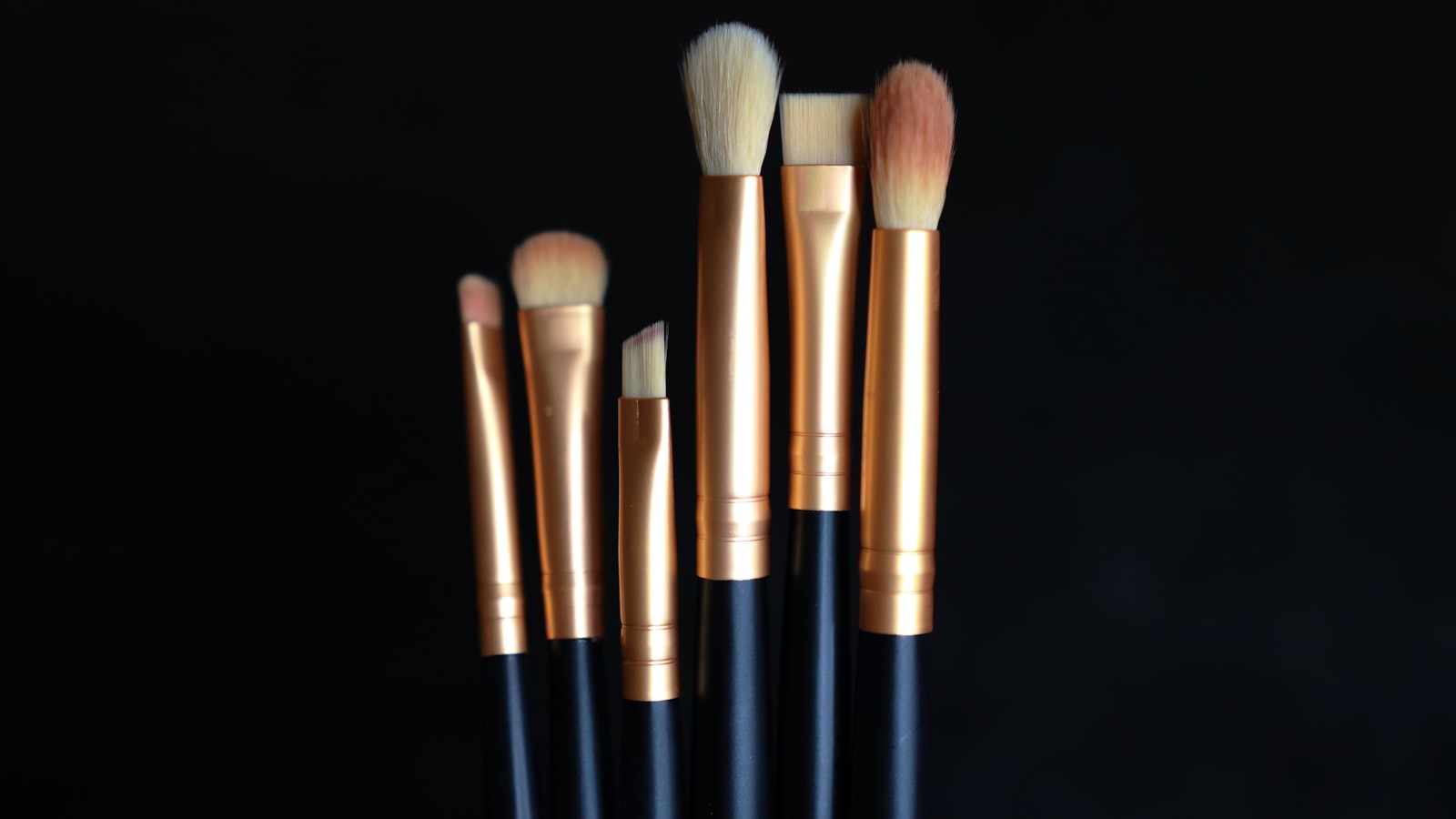 Six brushes with black handles and gold ferrules stand against a black background