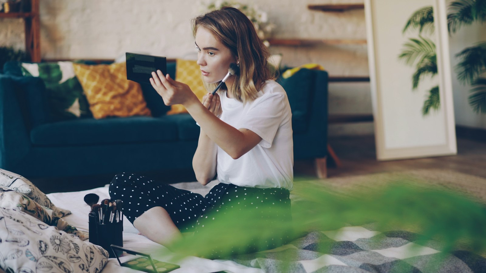 A young woman sits on the floor applying makeup with a brush, holding a small mirror in her other hand