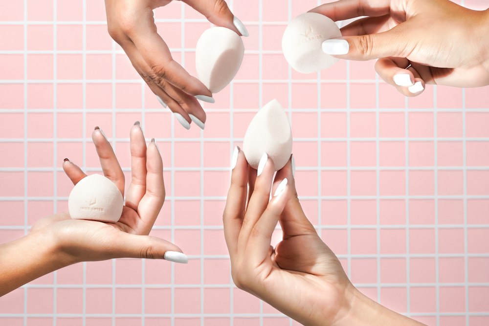 Four hands with white fingernails hold four white makeup sponges against pink tile