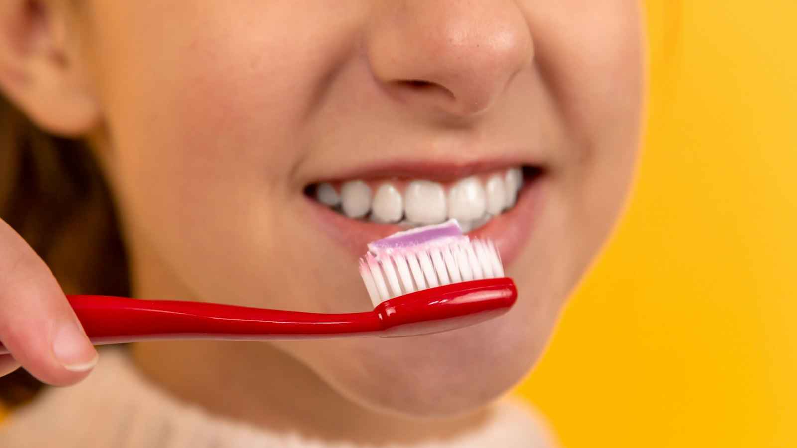 Close-up of a person holding a red toothbrush with toothpaste near their mouth against a yellow background