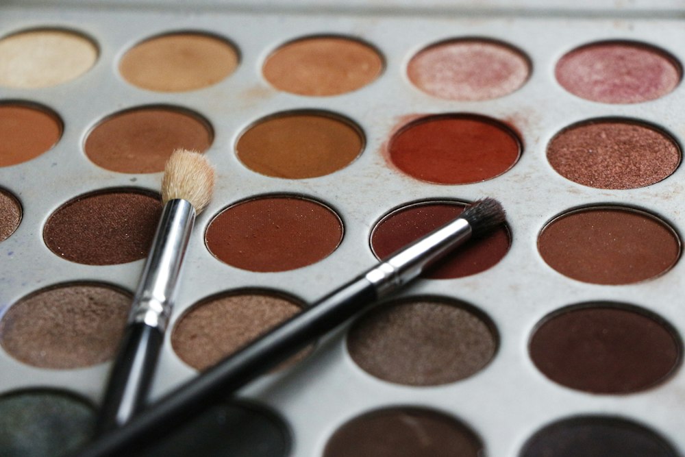 Two makeup brushes rest diagonally across an eyeshadow palette filled with circular pans of earth tones