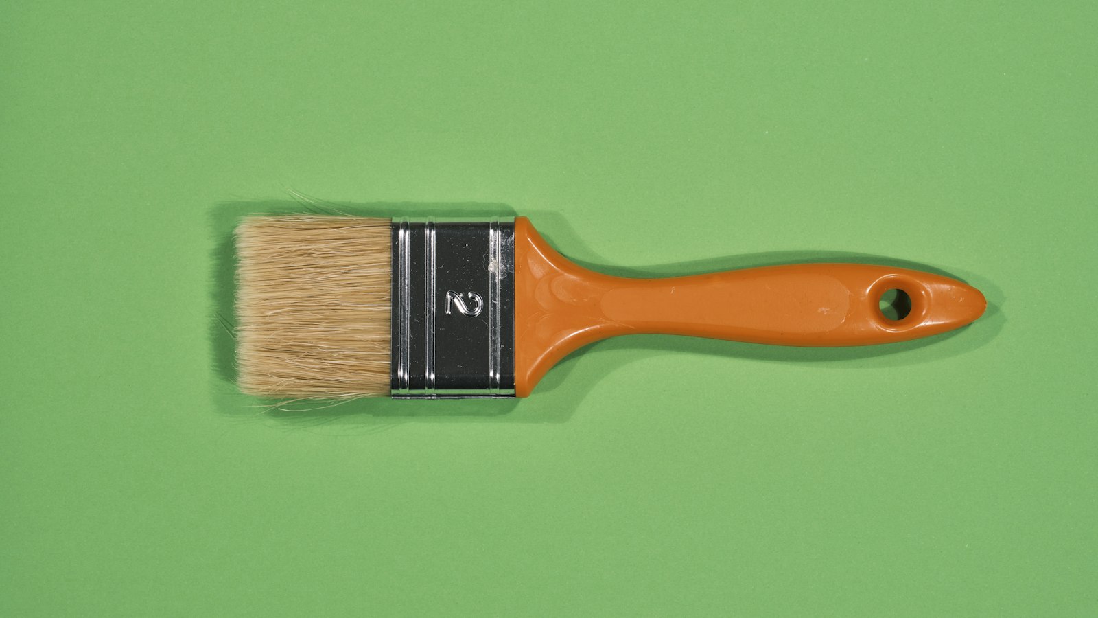 A paint brush with an orange handle and tan bristles rests on a green background