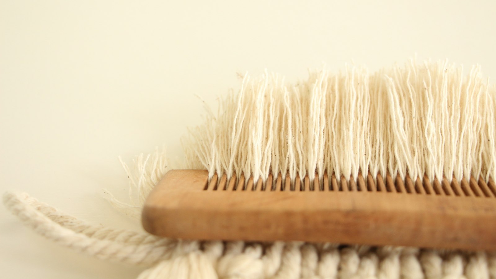 A wooden comb separating strands of off-white macrame cord against a plain background