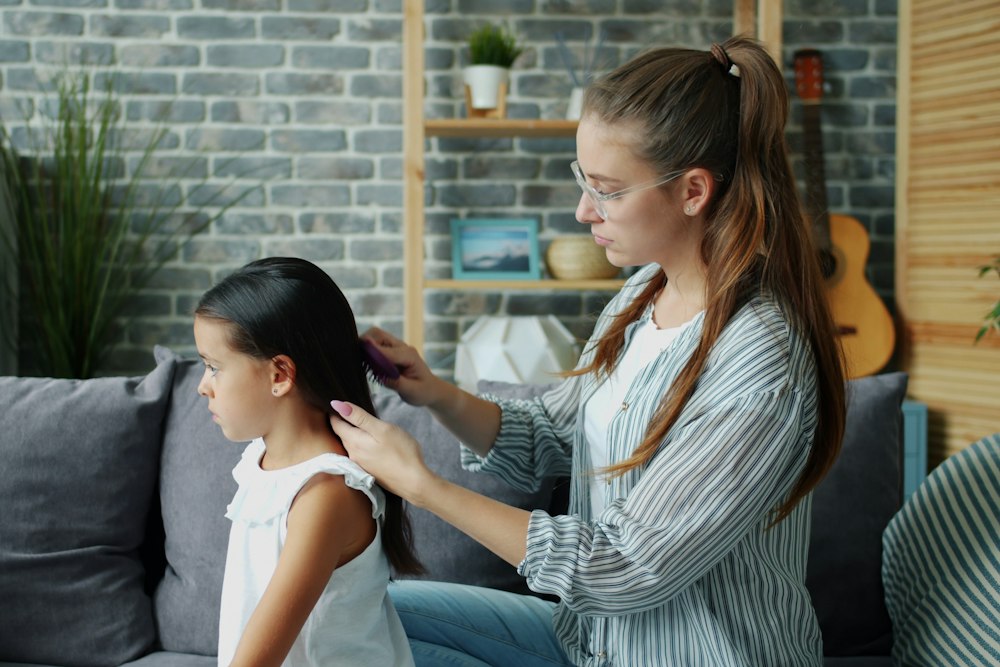 A woman in glasses combs the hair of a young girl sitting on a grey couch
