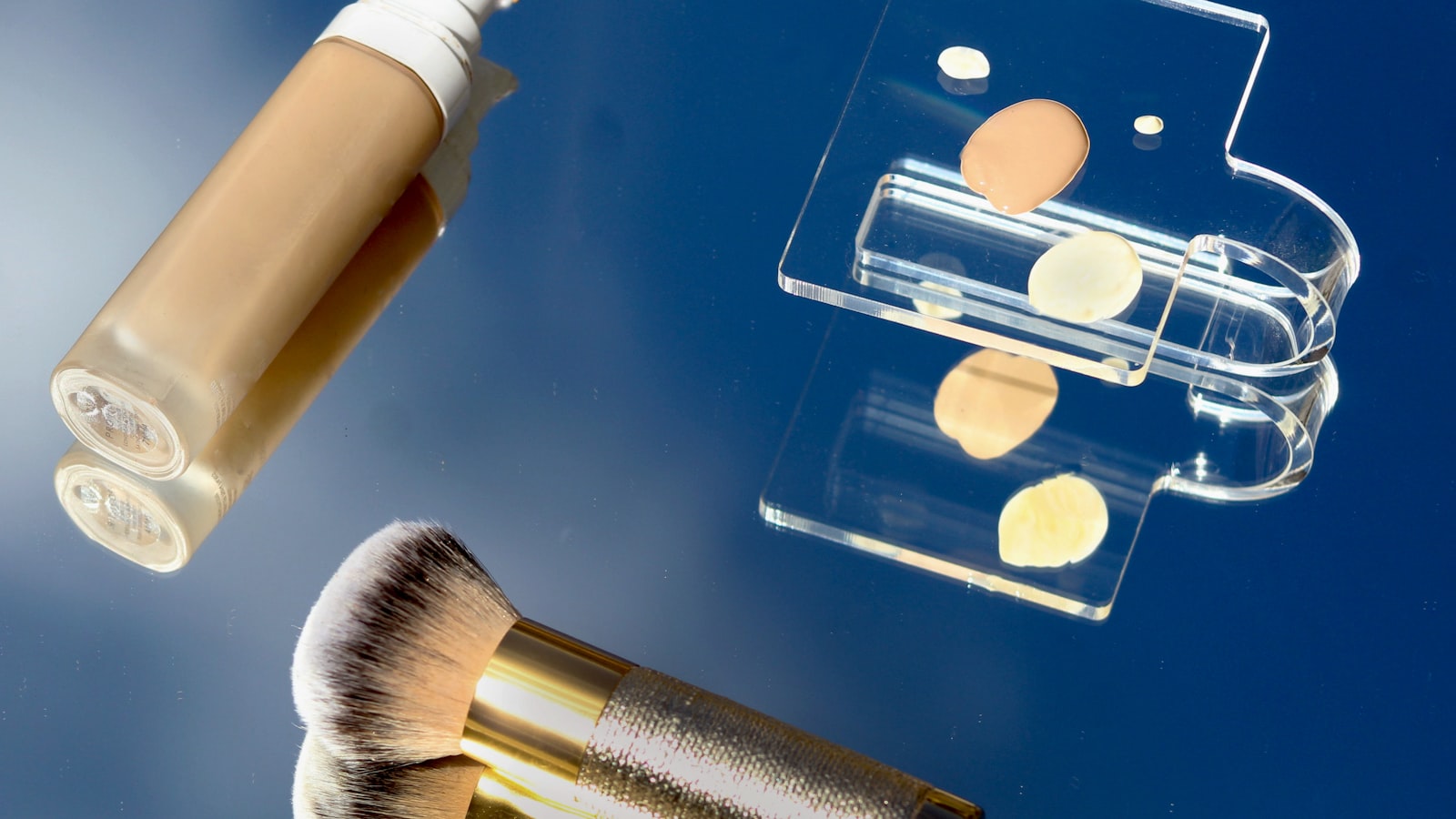 A bottle of foundation, a makeup brush, and a clear palette with dabs of cream on a reflective blue surface