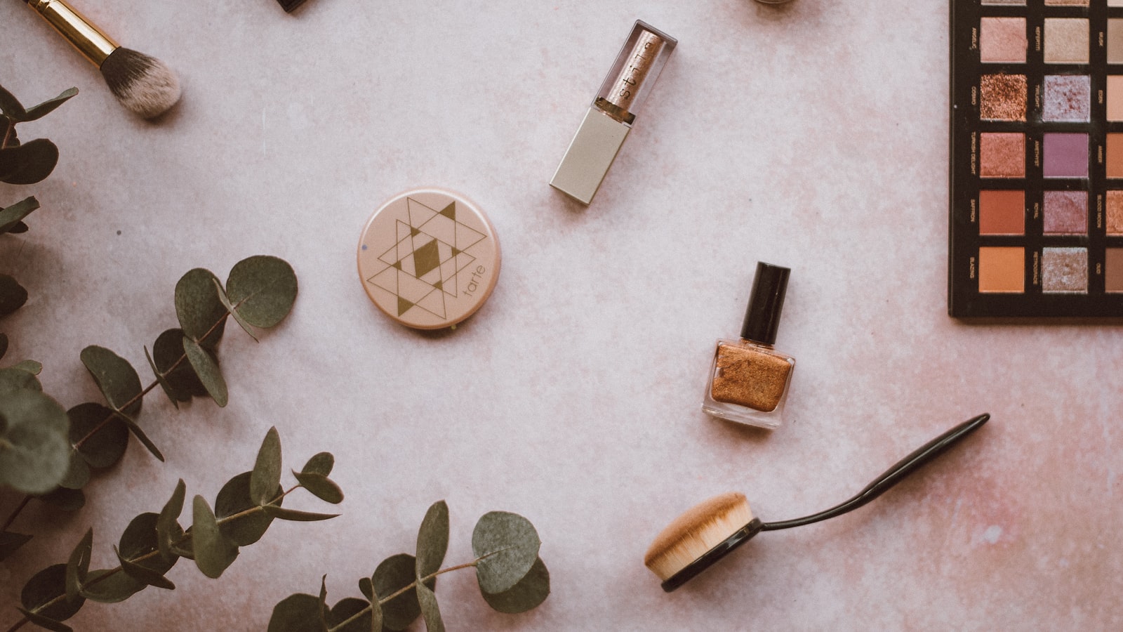 Makeup products and eucalyptus sprigs lie scattered across a pale pink surface