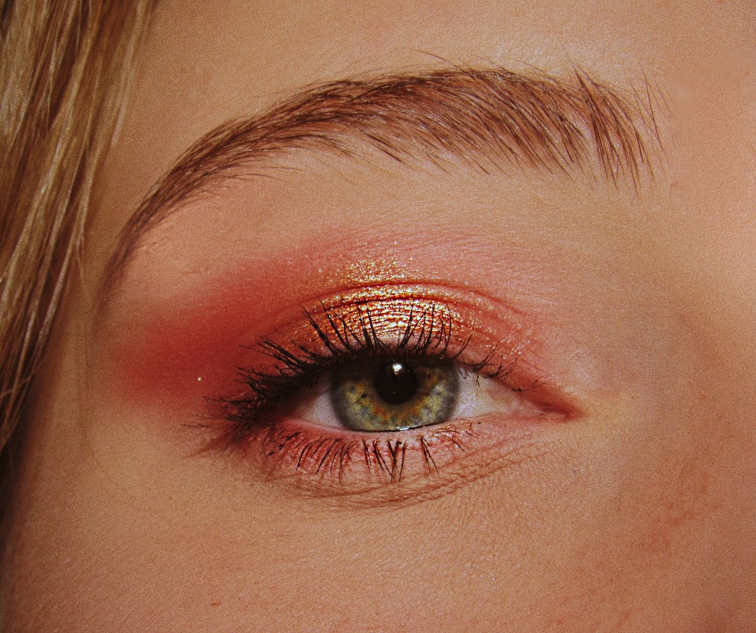 Close-up of a green eye wearing metallic gold and pink eyeshadow, with dark eyelashes and an arched eyebrow
