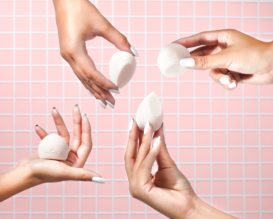 Four hands with white manicured nails hold four white makeup sponges against a pink tiled wall