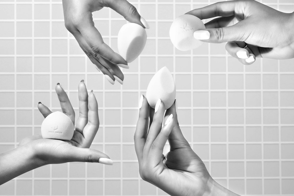 Four hands hold four different makeup sponges against a tiled wall in this black and white photo