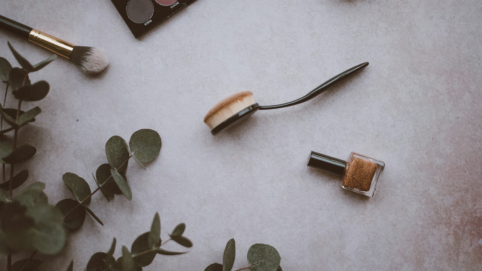 Overhead shot of makeup brushes, eyeshadow palette, and a small glass bottle on a grey surface with eucalyptus leaves
