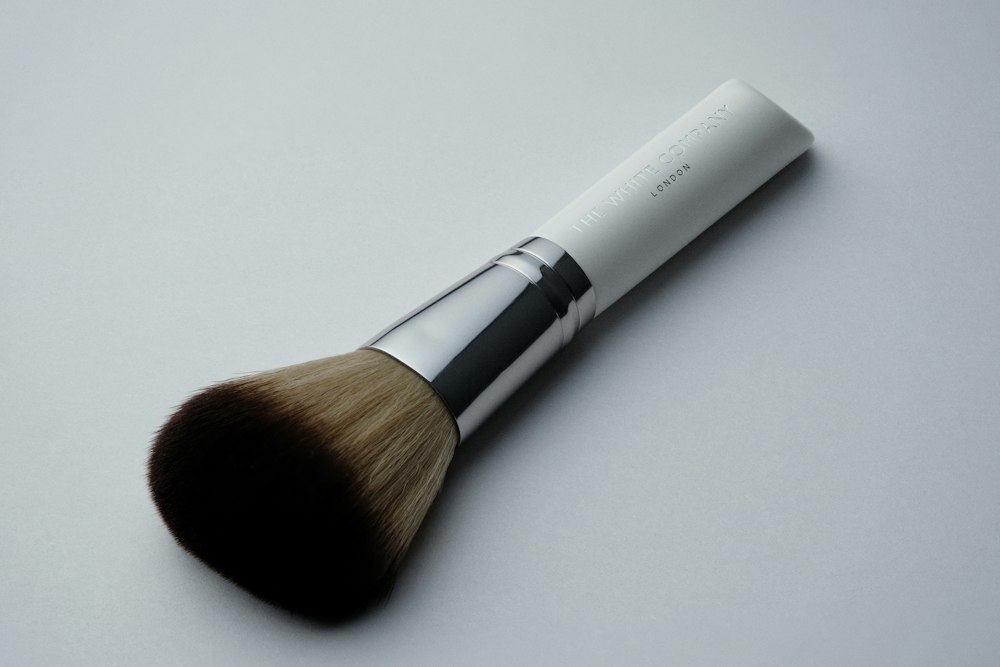 A cosmetic brush with a white handle, silver ferrule, and brown bristles lies diagonally on a grey surface