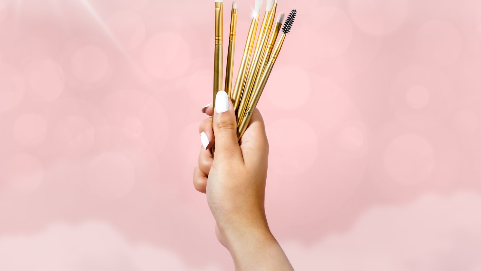 A hand with white fingernails holds a fan of gold-handled brushes against a pink background