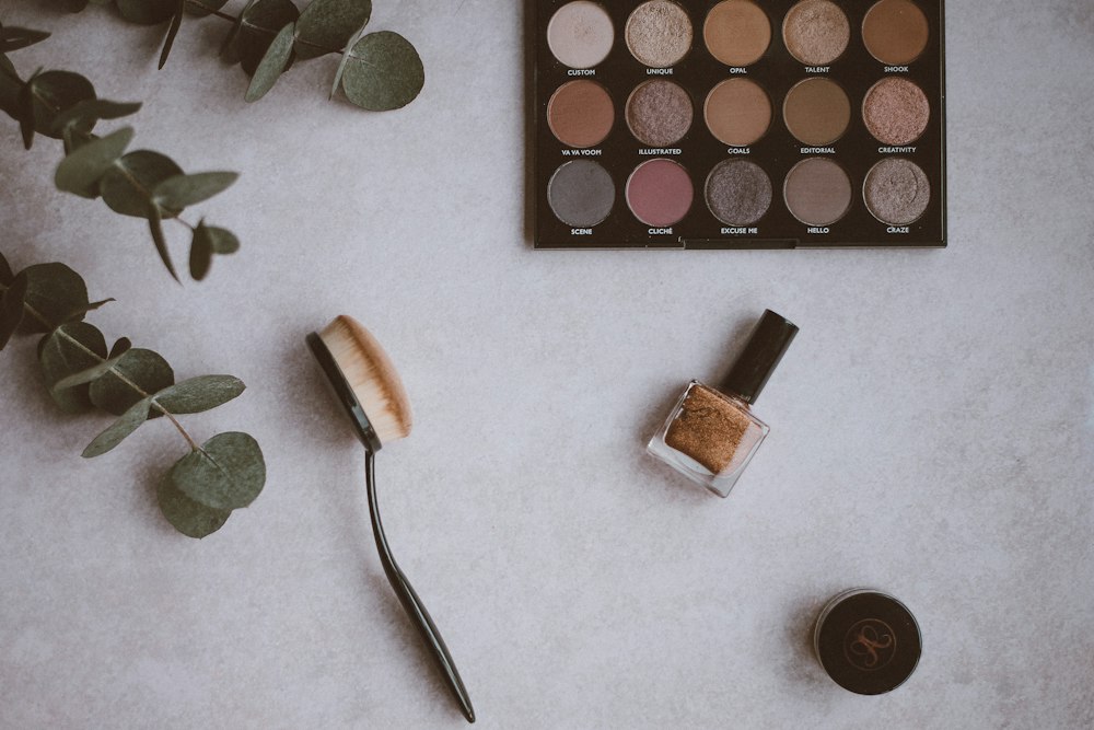 Eucalyptus branch, makeup palette, oval brush, nail polish bottle, and jar on a grey surface