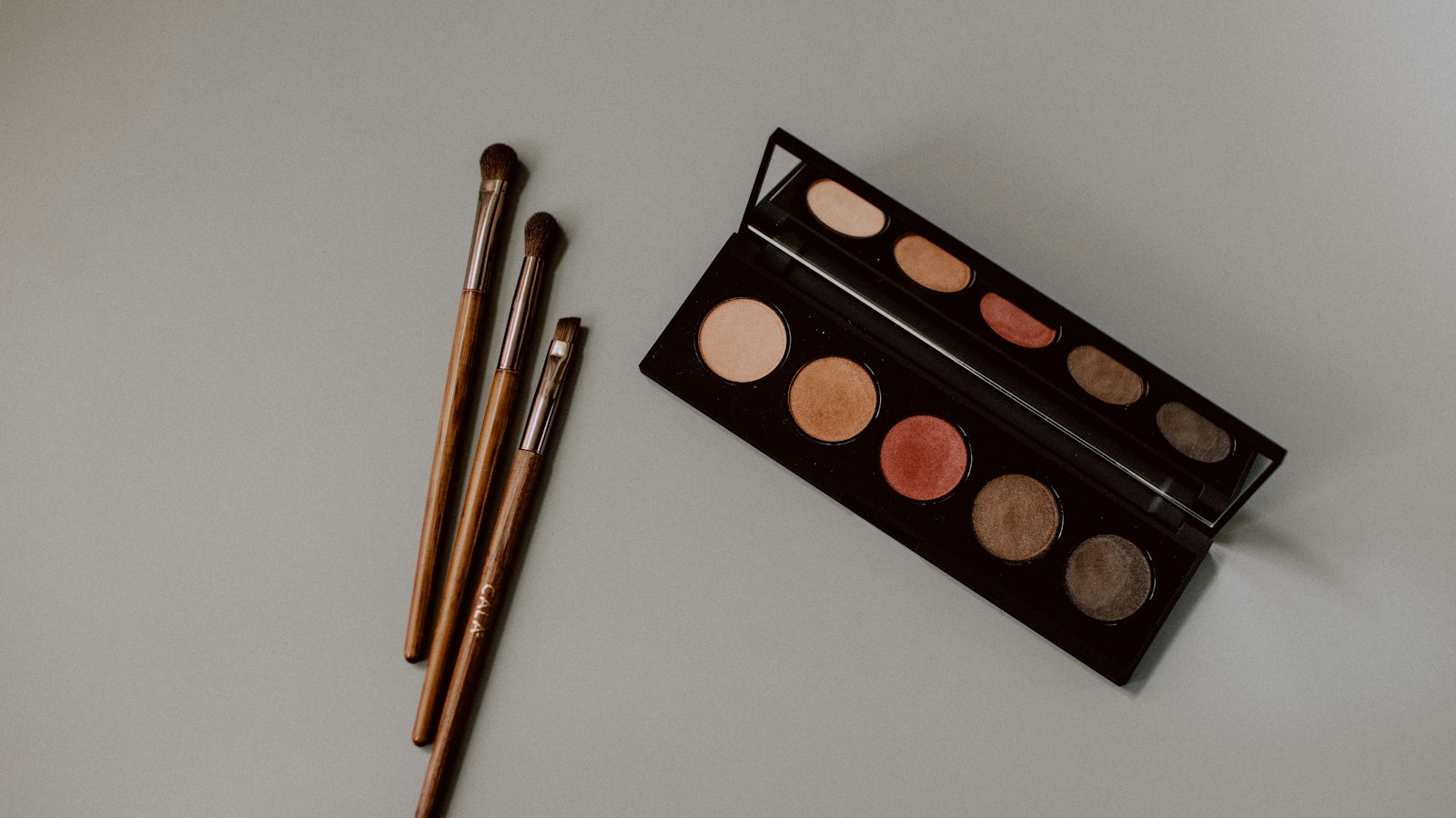 An open eyeshadow palette and three makeup brushes lie on a grey surface