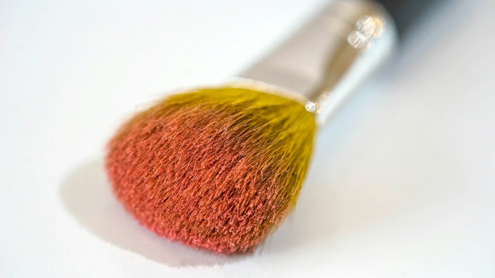 A close-up of a cosmetic brush with yellow bristles coated in pink powder against a white background