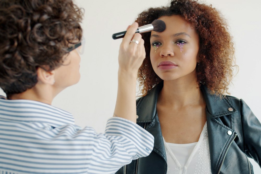 A woman in a striped shirt uses a brush on another woman's forehead
