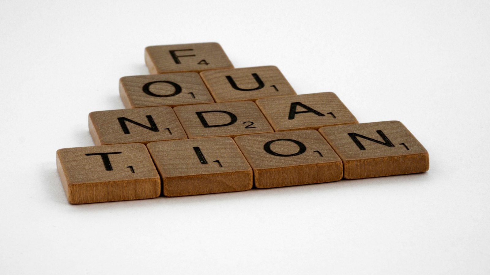 Wooden game tiles spelling the word "FOUNDATION" arranged in a pyramid on white