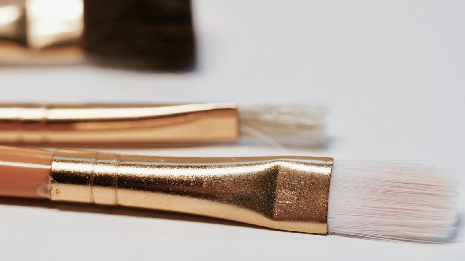 Three paintbrushes with gold ferrules and white bristles rest on a white surface