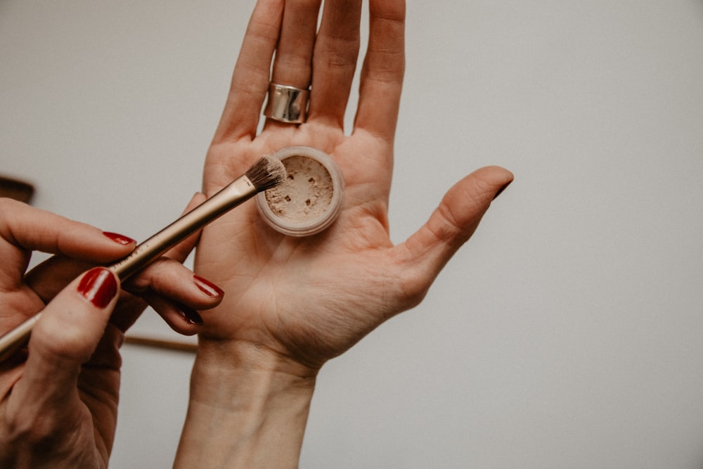 A person holds an open jar of powder and taps it with a makeup brush
