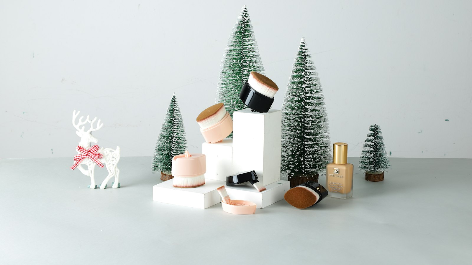 Brushes and a bottle of foundation are displayed on white blocks among miniature trees and a reindeer figurine