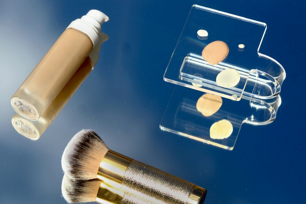 A bottle, a brush, and a clear palette with makeup drops sit on a reflective surface