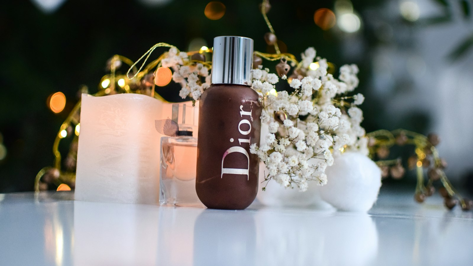 A bottle of Dior foundation sits on a table with a candle and small white flowers