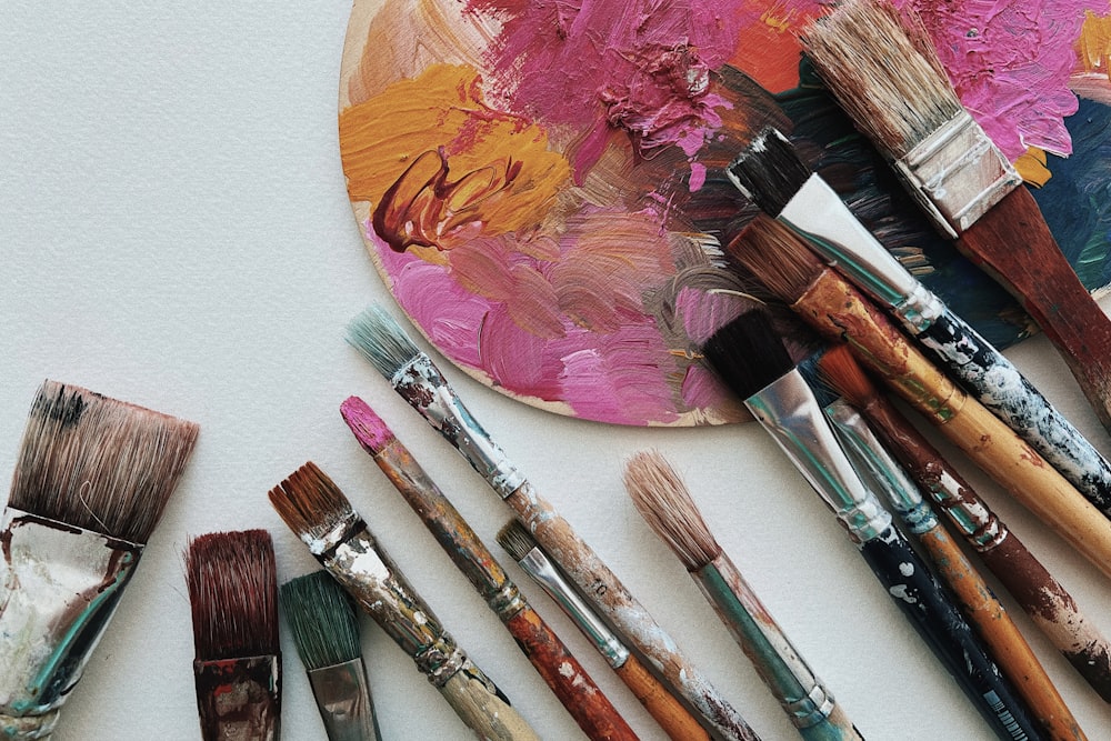 An art palette with pink and orange paint is surrounded by various paintbrushes