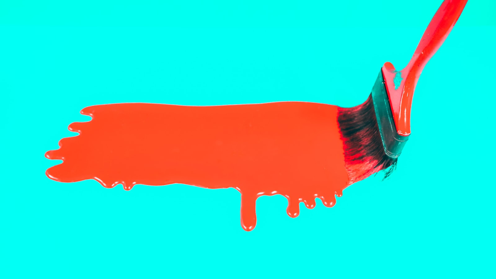A paintbrush with red paint strokes a thick red stripe horizontally across a bright turquoise surface