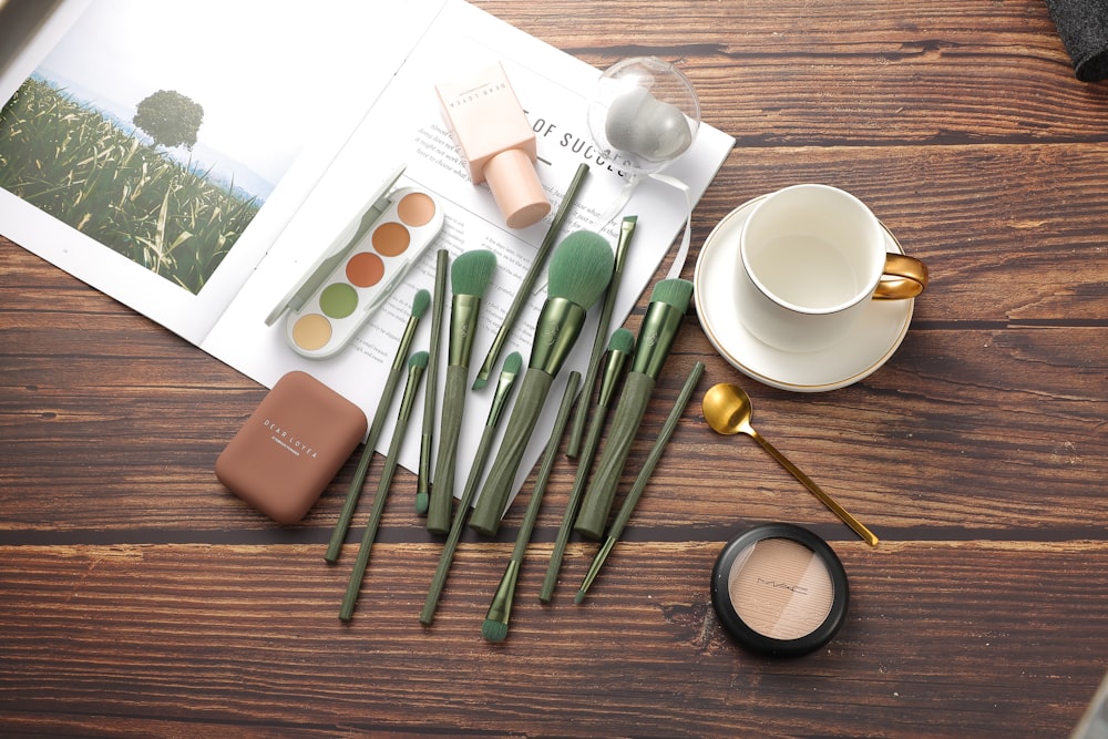 A flat lay of makeup brushes, palettes, a cup, and a magazine on a wooden surface
