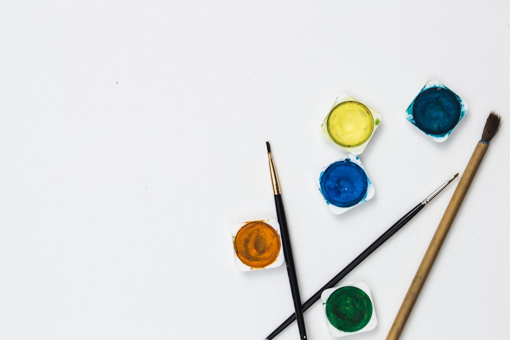 Top-down view of five watercolor pans in yellow, blue, orange, green, and dark blue, with three paintbrushes on white paper